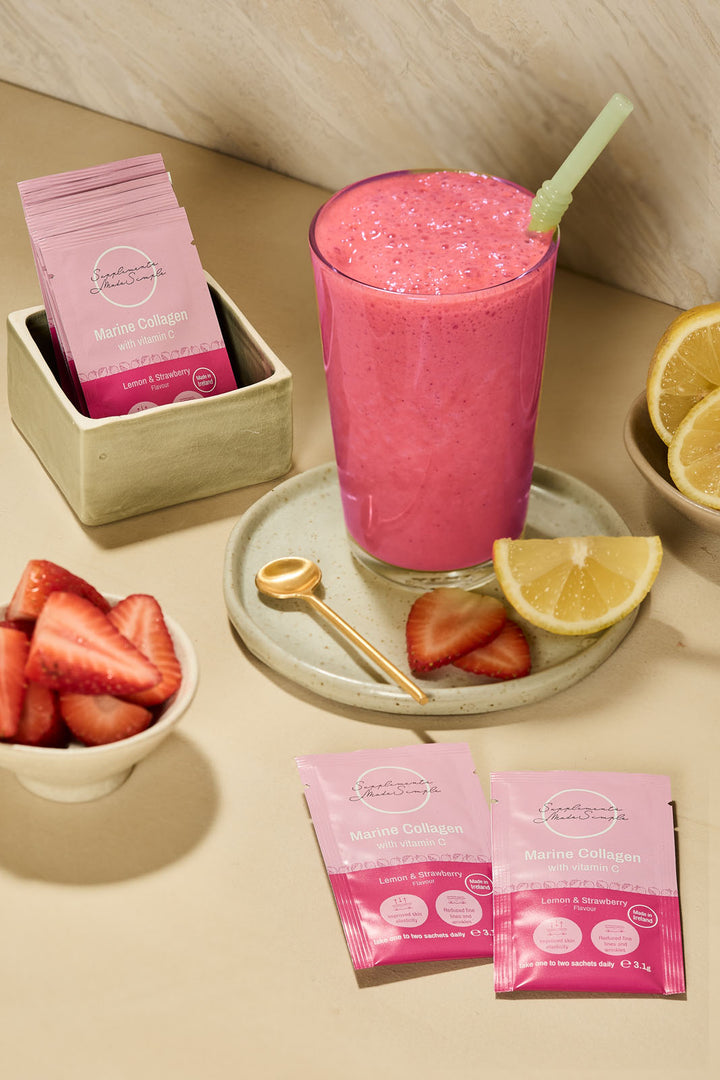 A pink smoothie in a tall glass with a straw sits on a tray beside two pink sachets of protein powder, with bowls of strawberries, lemon slices, and a small spoon on a beige tabletop.